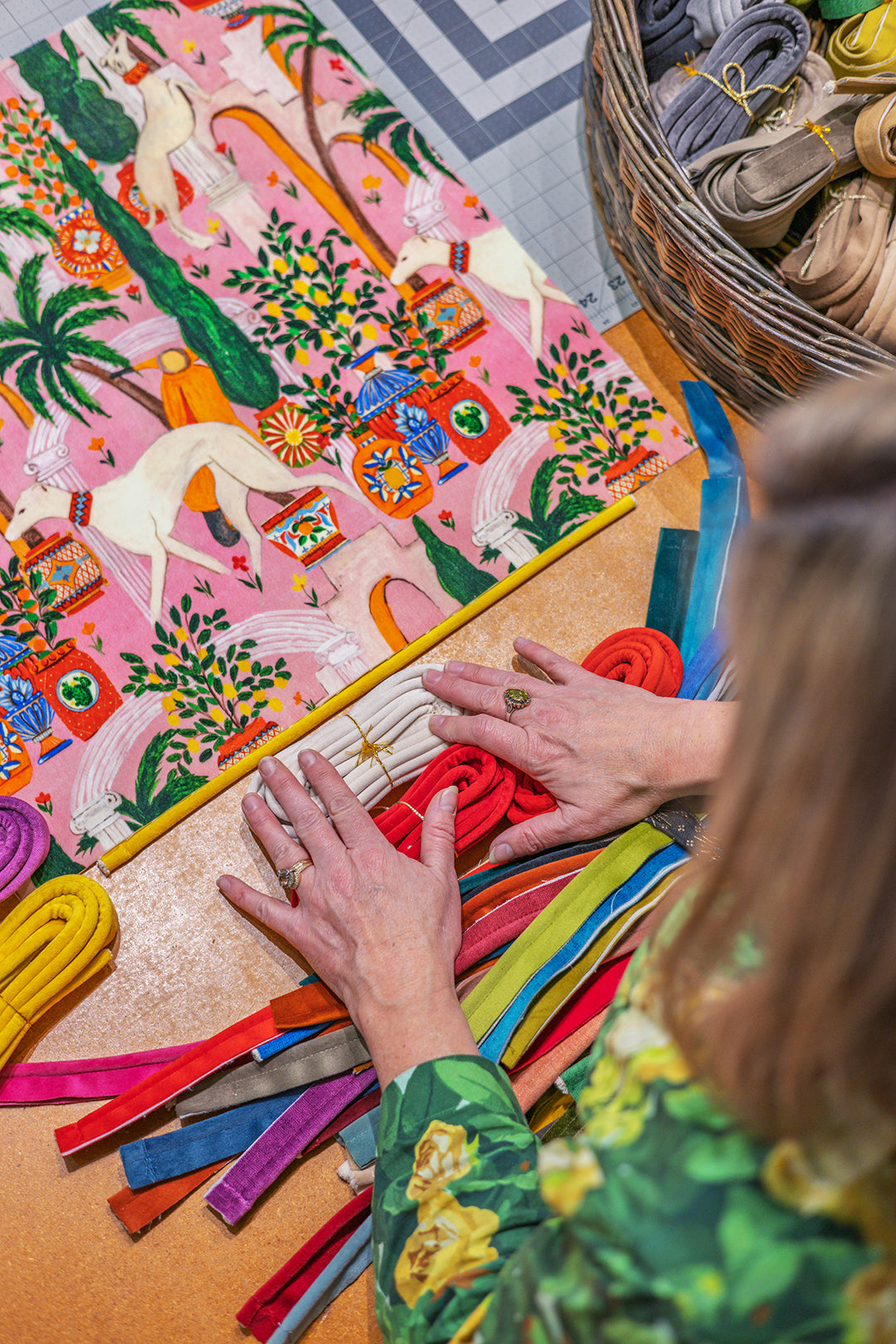 A close-up overhead view of a person's hands arranging colorful fabric strips on a wooden table next to a large pink patterned textile. The scene includes a basket of rolled fabrics in the background, with the person wearing a green floral top.