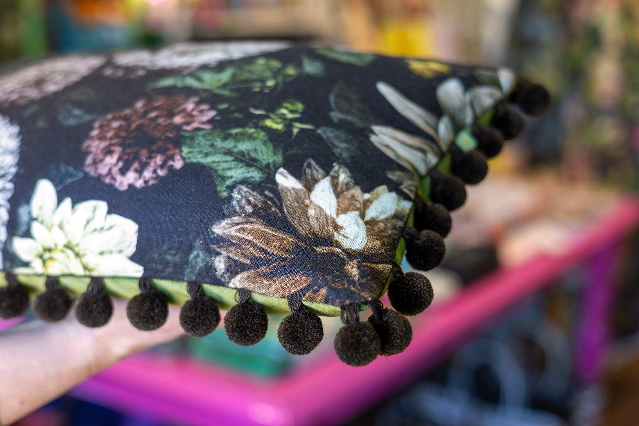 A close-up of a decorative throw pillow with a black background and a multicolor floral print, finished with dark brown pom-pom trim along the edge. A hand is holding the pillow in a brightly lit, blurred shop setting.