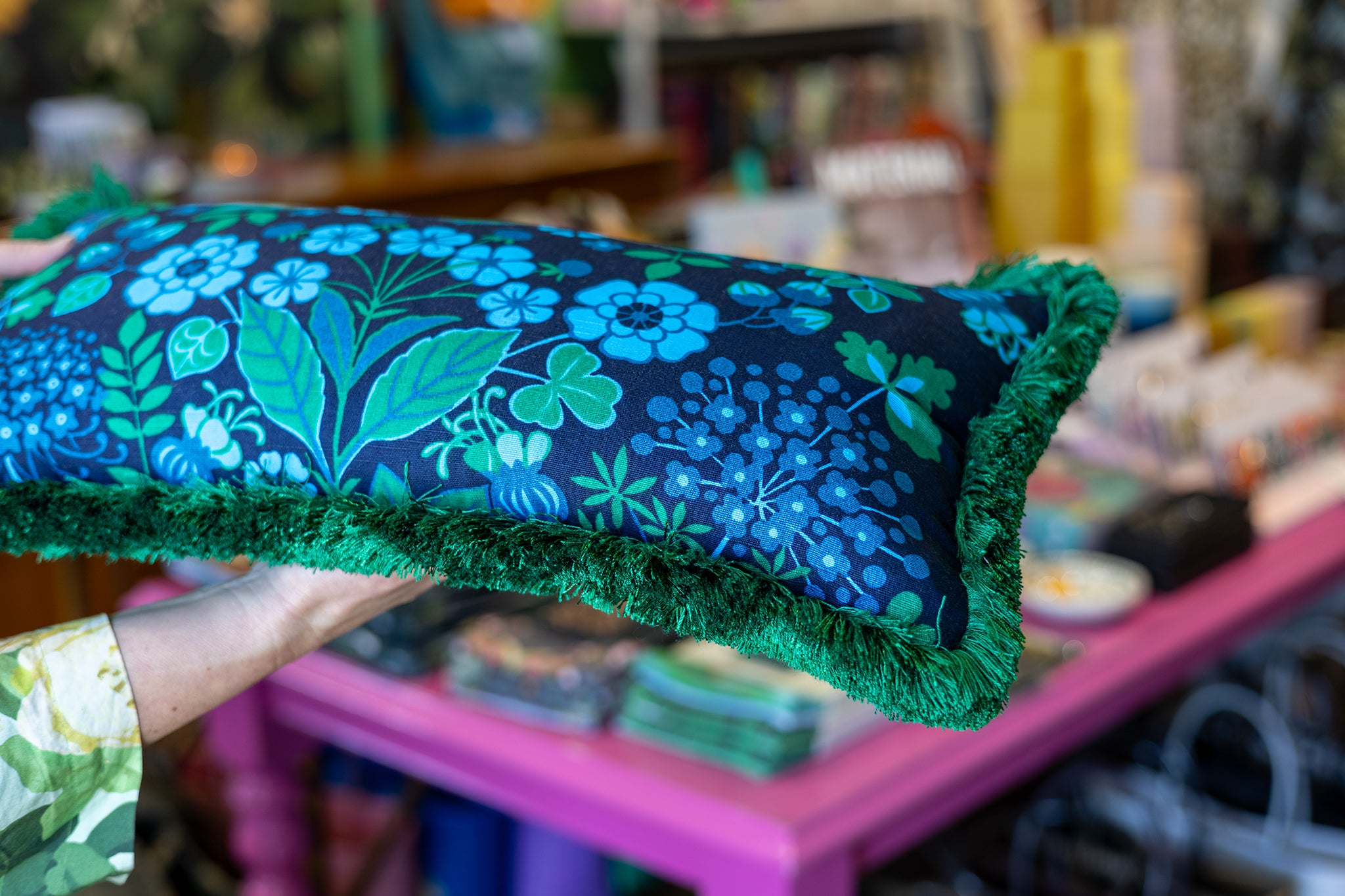 A hand holds a rectangular decorative pillow with a dark navy blue background, a vibrant blue and green floral pattern, and a thick green fringe trim, set against a blurred shop background.