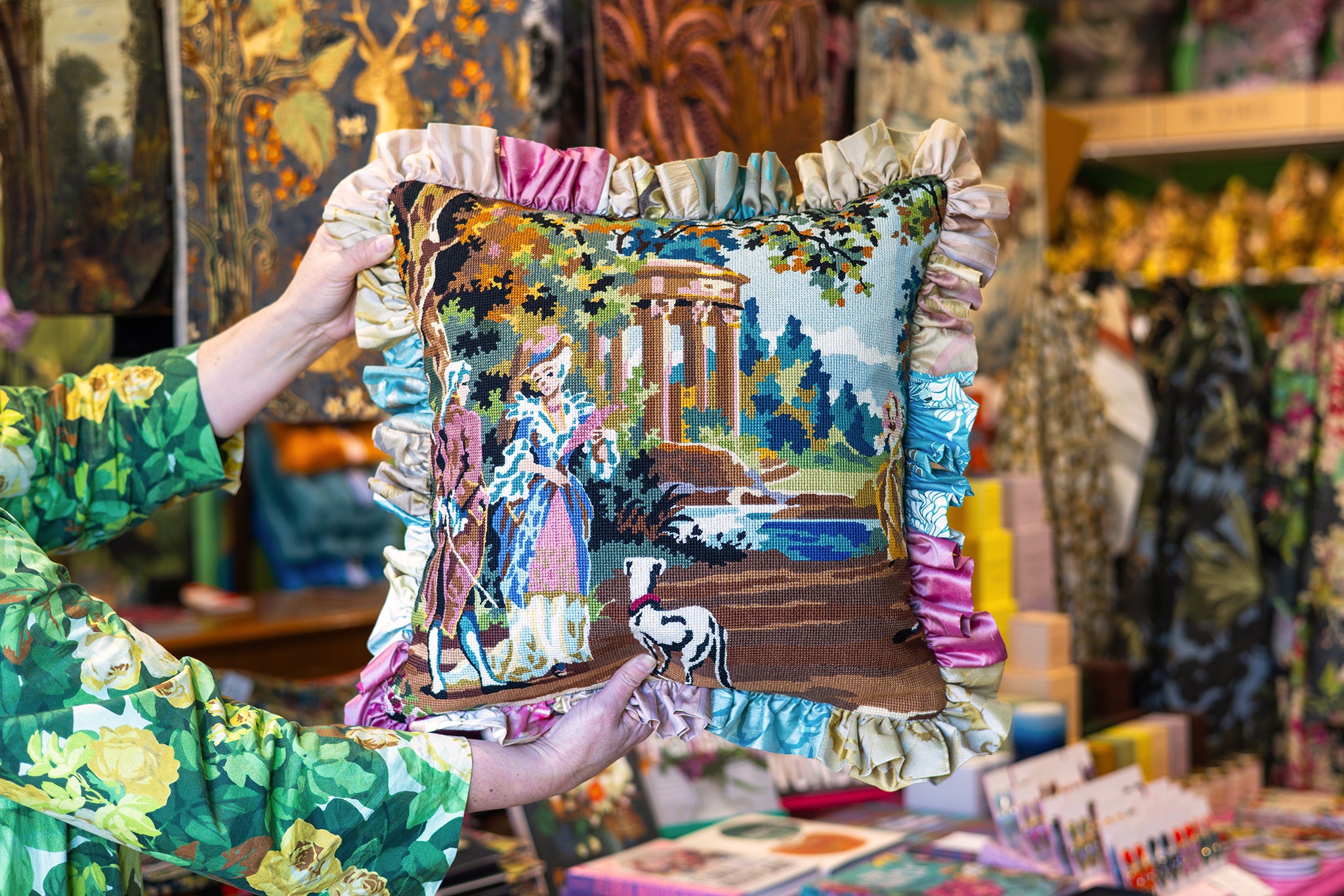 A person holds up a square decorative pillow in a shop. The pillow features a colorful embroidered scene of a woman in a classical landscape with a river and trees, surrounded by a ruffled trim in pastel shades of blue, pink, and beige.