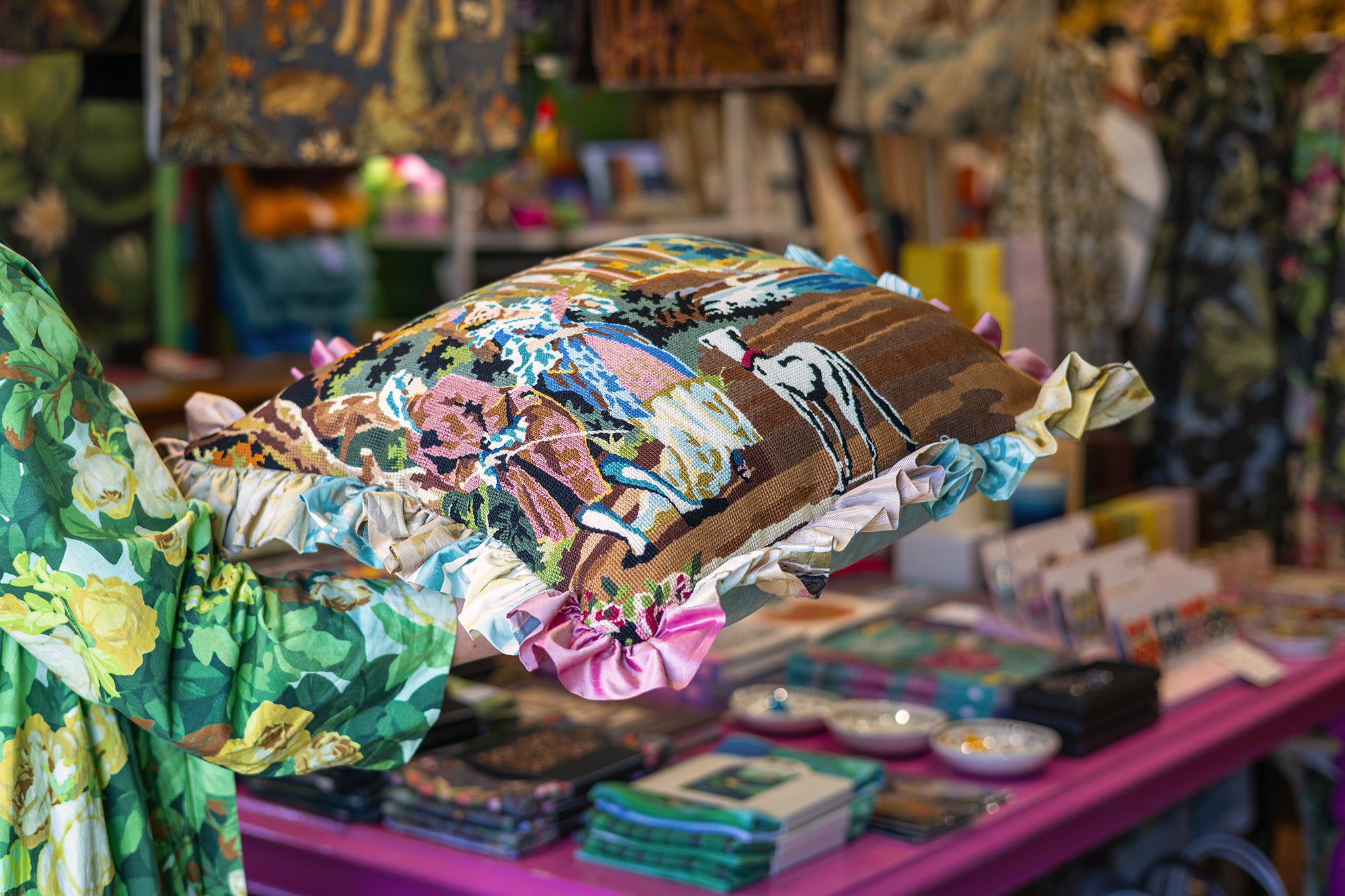A person in a green floral dress holds up a large tapestry pillow in a shop with a pink table and other textiles in the background.
