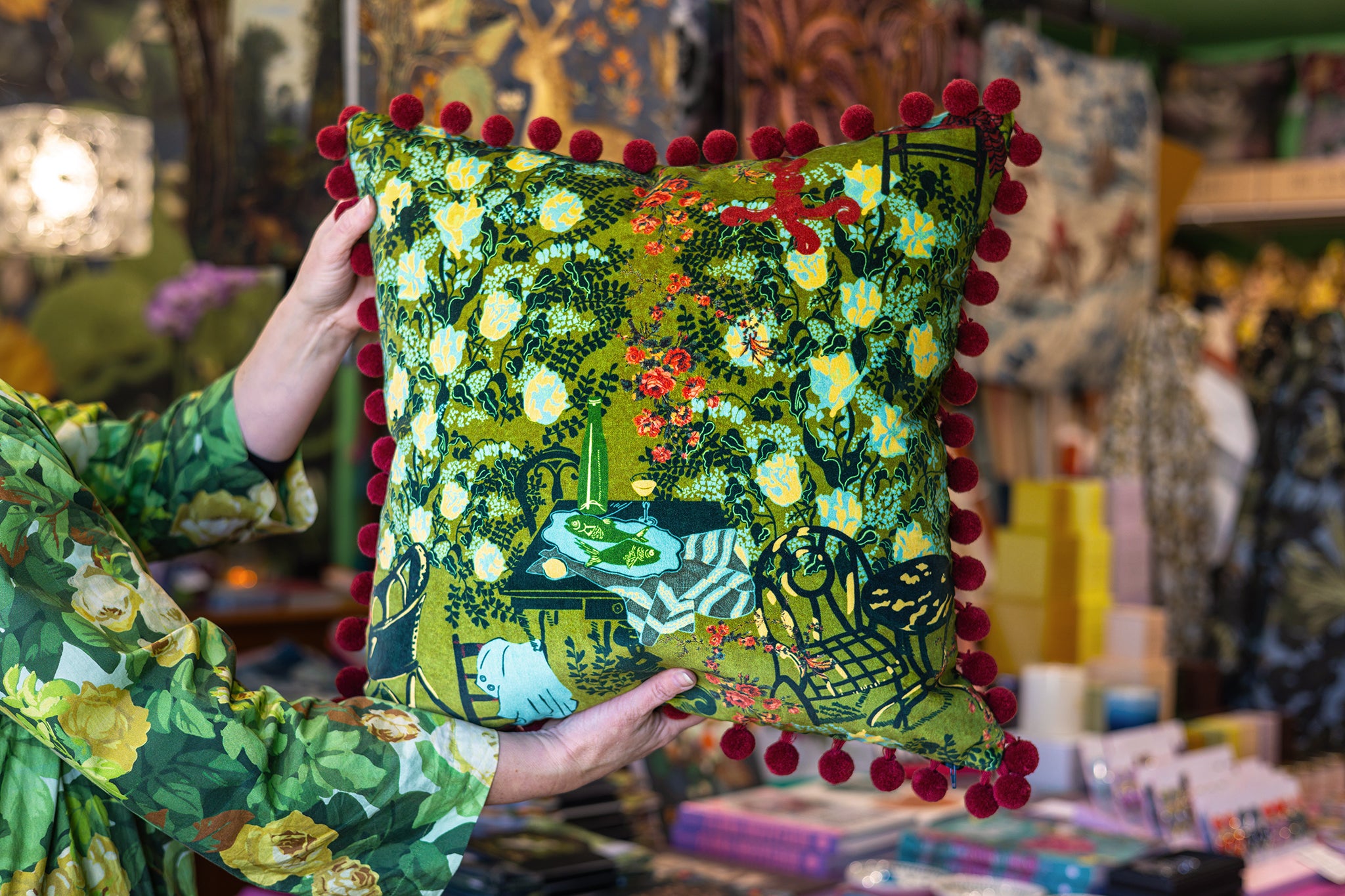 A person holds up a square decorative throw pillow with a green and yellow floral print and red pom-pom trim, set against a busy, colorful shop background.