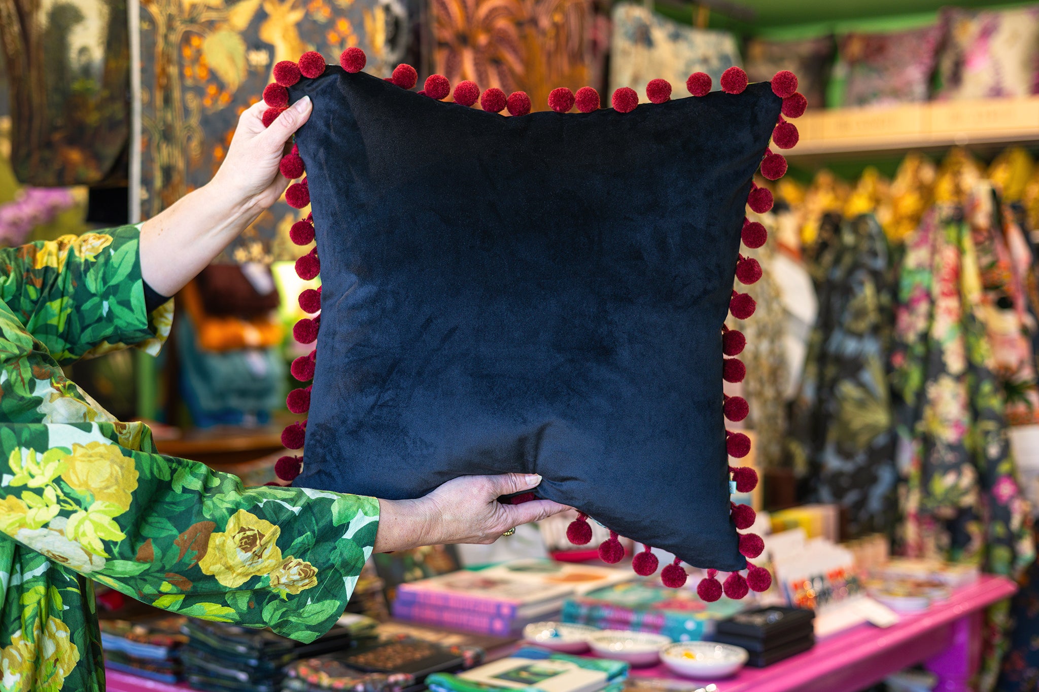 A person holds up the black velvet back of a square cushion framed with pom poms in a busy shop
