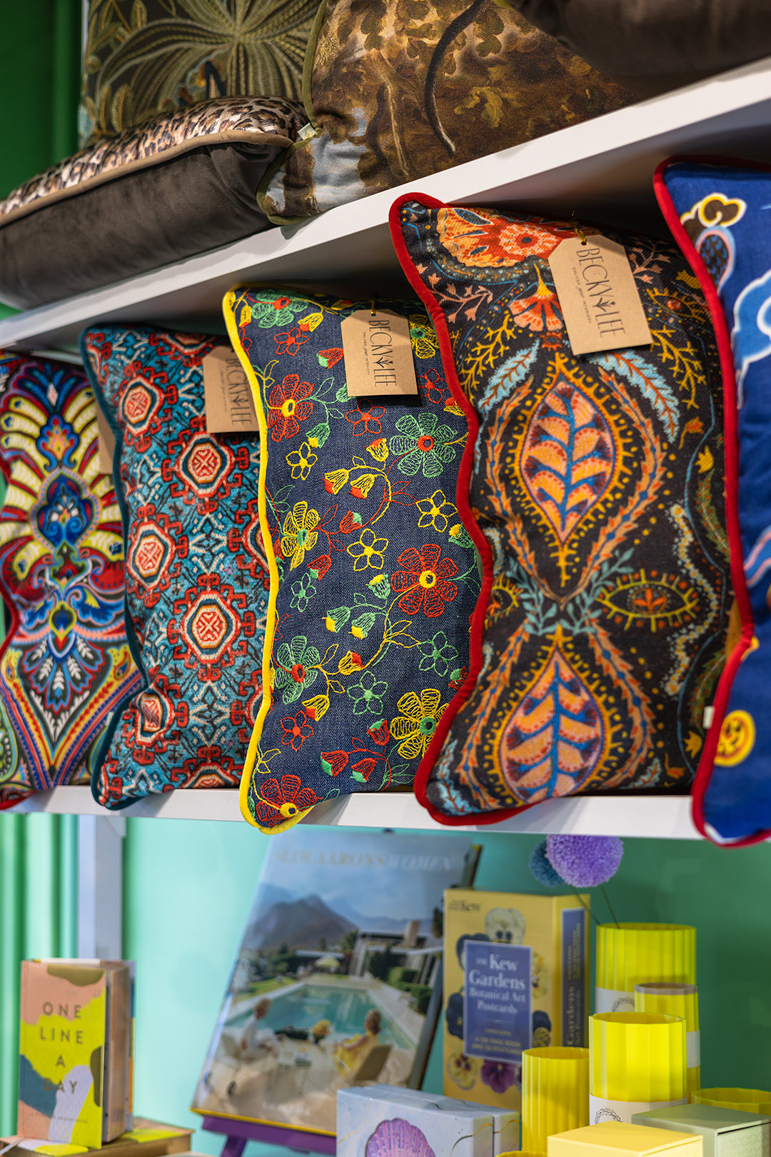 A close-up of several colorful decorative throw pillows displayed on a white shelf. The pillows feature intricate hand-woven or printed patterns in blue, red, yellow, green, and orange, with some showing red piping.