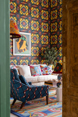 Vibrant lounge with bold Amalfi-style wallpaper, a white sofa with patterned cushions, a blue armchair, colourful rug, and eclectic décor. POV is doorway of room peeking in