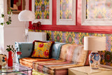 Colourful lounge with a mix of patterned textiles, a glass coffee table, vibrant Amalfi-style wallpaper, a white lamp, and eclectic décor accents.