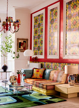 Colourful eclectic living room with patterned Amalfi wallpaper, a vibrant patchwork sofa, bold rug, red chandelier, and artistic decor.