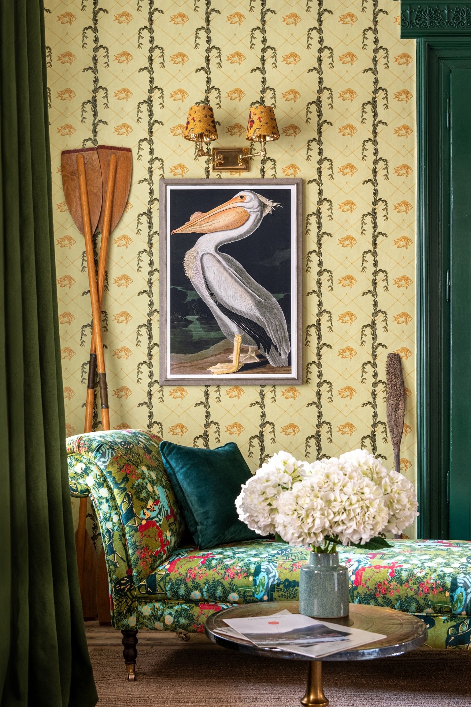 A cozy room with green floral wallpaper, a vintage chaise lounge with a green velvet pillow, oars, and a framed pelican print