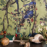 Close up of BIRDS OF DANUBE wallpaper with vibrant blue jays on branches, set behind eclectic decor—sculptures, antique books, and rustic ornaments