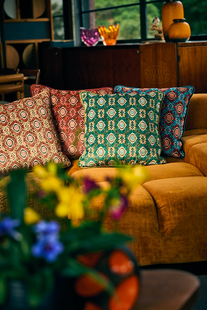 Four decorative throw pillows with intricate geometric and floral patterns in red, green, blue, and beige, arranged on a mustard yellow fabric sofa in a cozy, softly lit living room setting.