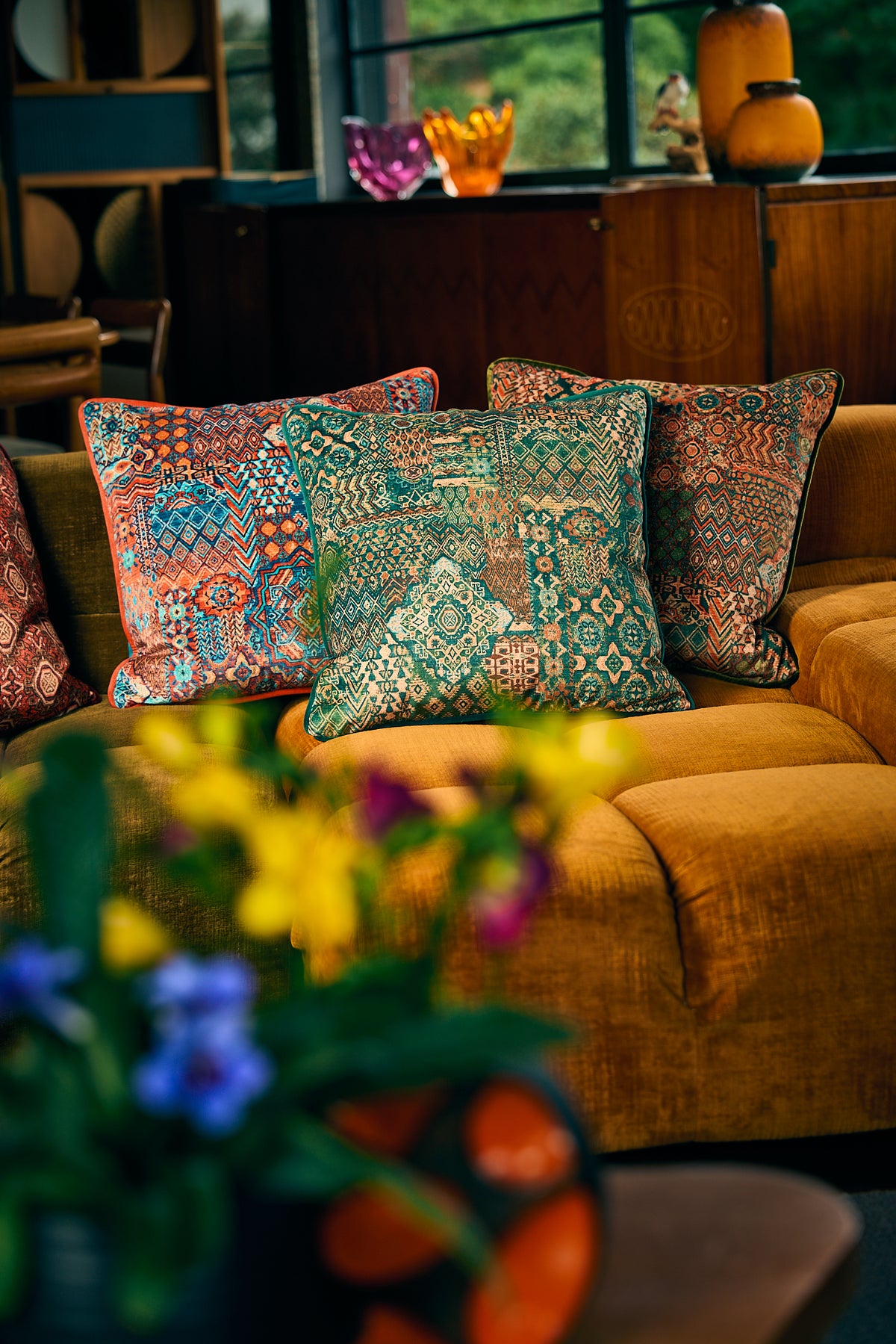 A cozy living room scene featuring a mustard yellow velvet sofa adorned with three decorative throw pillows in intricate teal, rust, and orange patterns. In the foreground, a blurred vase of colorful flowers adds a vibrant touch, while a dark wood cabinet and window are softly visible in the background.