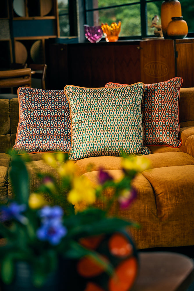 Three decorative throw pillows with geometric patterns in red, orange, and green tones, arranged on a mustard yellow sofa in a cozy living room setting with blurred flowers in the foreground.