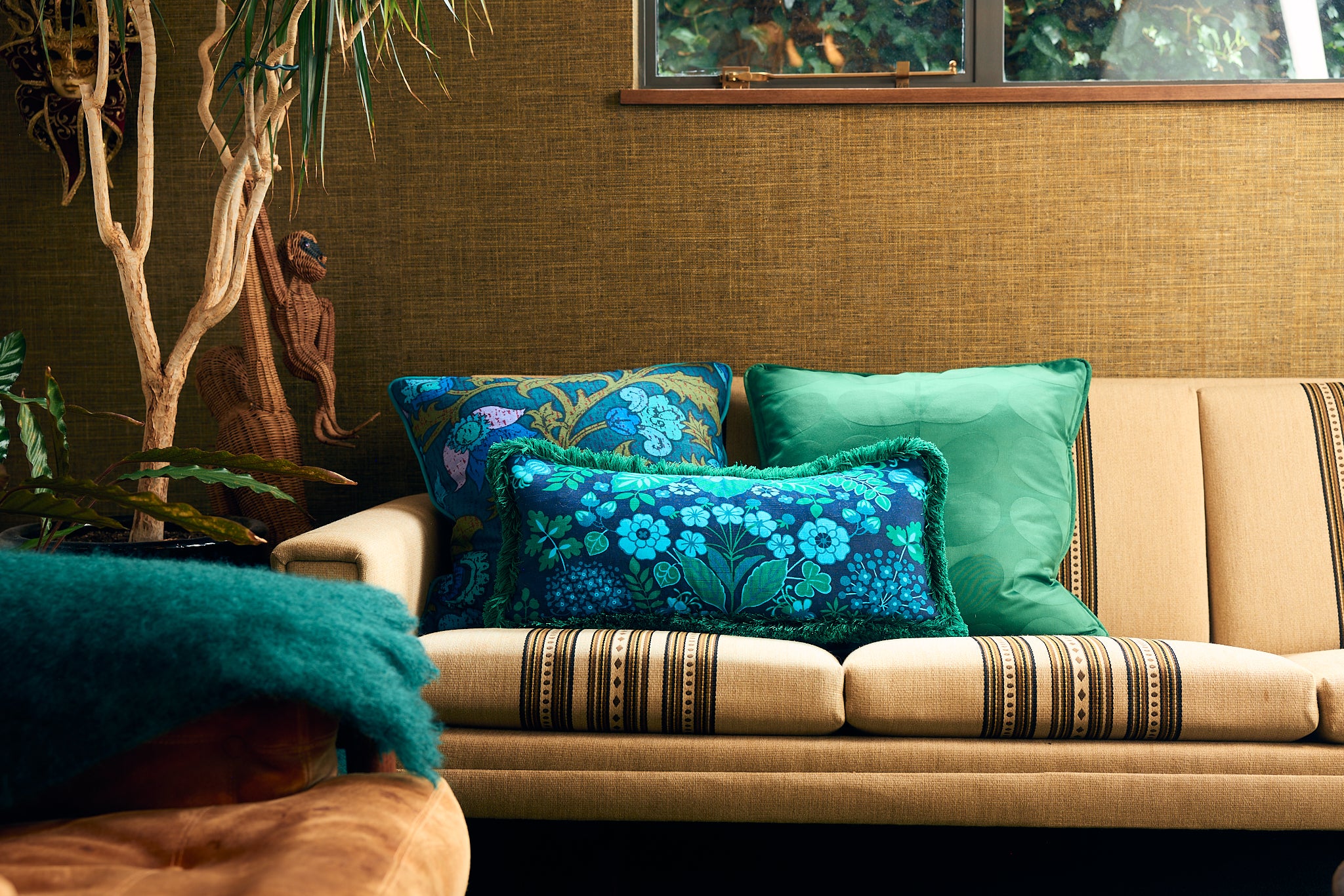 A styled interior scene showing a vintage upholstered sofa with decorative throw pillows in teal, turquoise, and blue floral patterns, set against a textured olive-brown wall with a window and indoor plant visible.