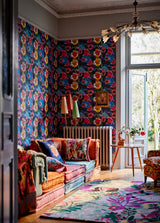 Cozy living space with bold floral FIORI IN FIORE wallpaper, a layered cushion sofa, colourful textiles, vintage lighting, and an open window with garden views.