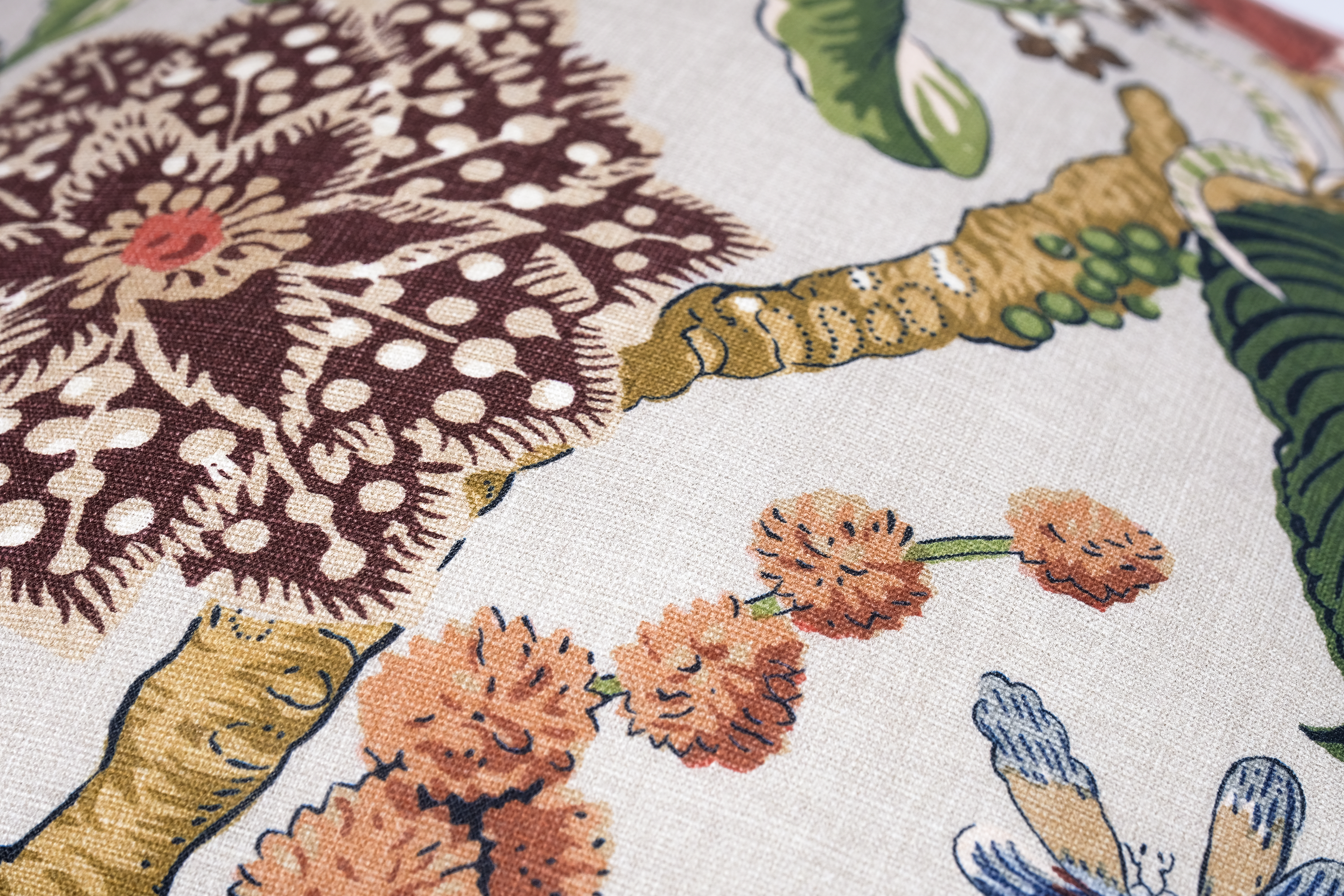 A close-up of a woven fabric with a detailed botanical pattern featuring brown, beige, green, and orange elements on a light cream background.