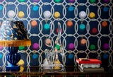 Luxurious interior with bold geometric INCATENATO Blu wallpaper featuring colourful spheres, a fringed leopard-print lamp, crystal sculpture, and stacked books.