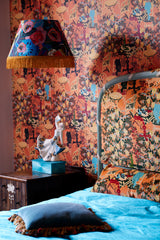 Bedroom with LA BRASSERIA Salmon wallpaper featuring vintage café scenes, a matching upholstered bed, blue bedding, a fringed lamp, and antique decor.