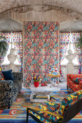 Eclectic living space with LE DONNE DI GIOIA wallpaper, bold patterned furniture, classical busts, vibrant textiles, and ornate decor. Concrete arched ceilings