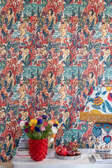 Bold LE DONNE DI GIOIA wallpaper featuring nude figures and lush botanicals, complemented by a red vase with colorful flowers, a fringed lamp, and fresh pomegranates.