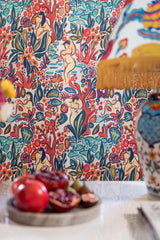 Close-up of LE DONNE DI GIOIA wallpaper with abstract nude figures, a fringed lamp, and a table with fresh pomegranates and decorative accents.
