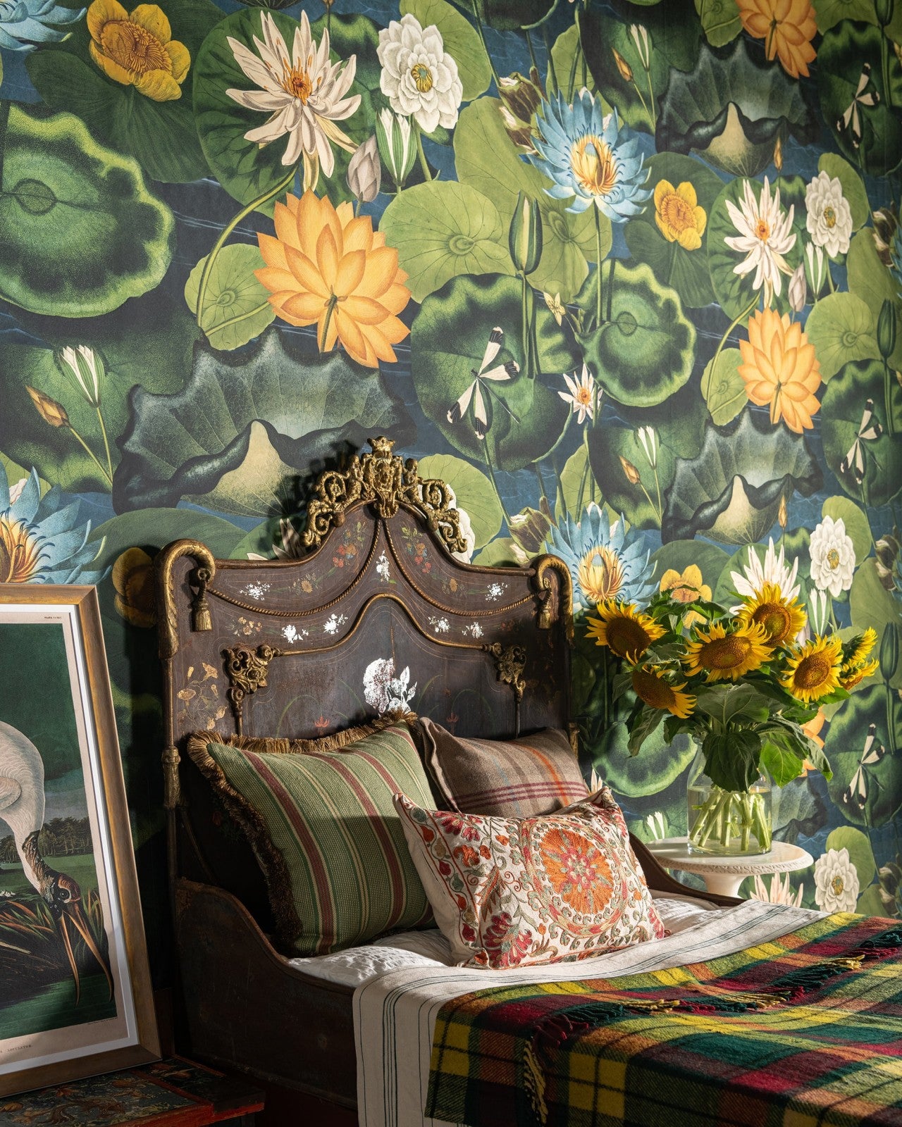 A vintage-style bedroom with an intricately carved wooden bed, floral-patterned pillows, and bold water lily wallpaper. Sunflowers in a vase bring warmth to the space