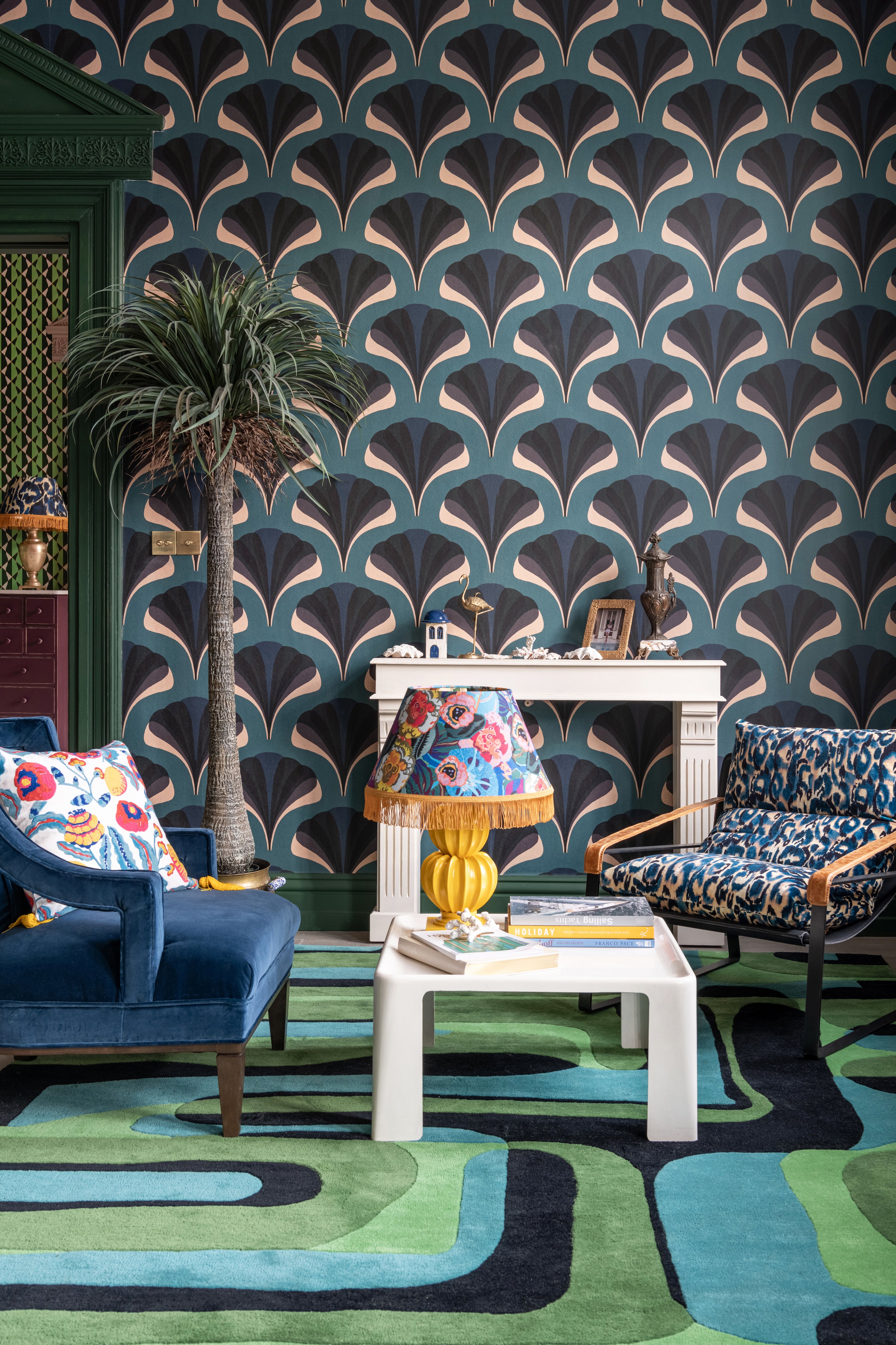 Sophisticated living space with MEZZELUNE Kale wallpaper, a bold geometric rug, velvet armchairs, a fringed lamp, and an ornate fireplace.