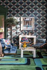 Elegant living room with MEZZELUNE Kale wallpaper, a bold green and blue rug, velvet armchairs, a fringed lamp, and a stylish faux fireplace.