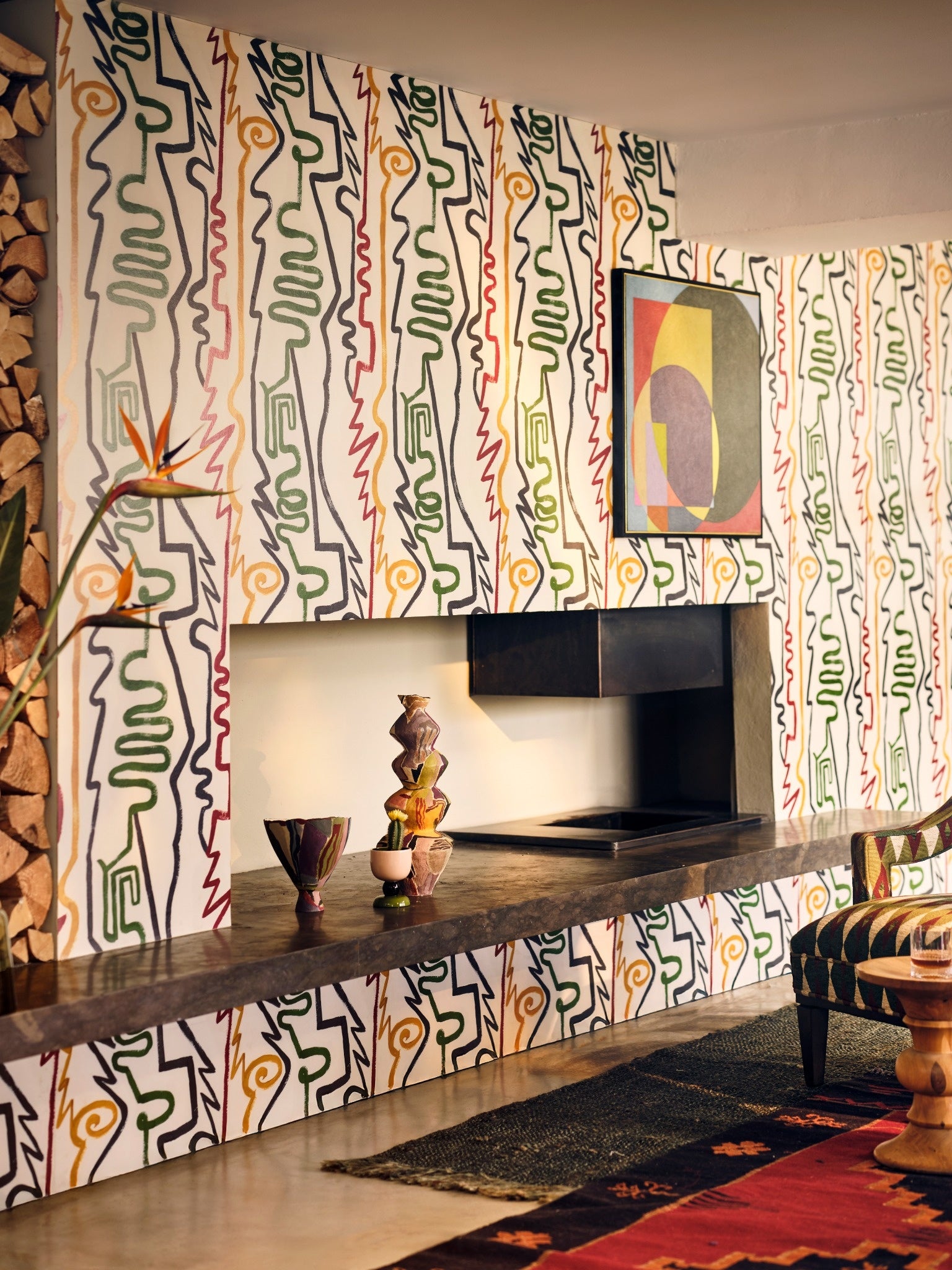 Colourful abstract line-drawing wallpaper with green, yellow, red shapes surrounding a black stone shelf, vases, and modern artwork.