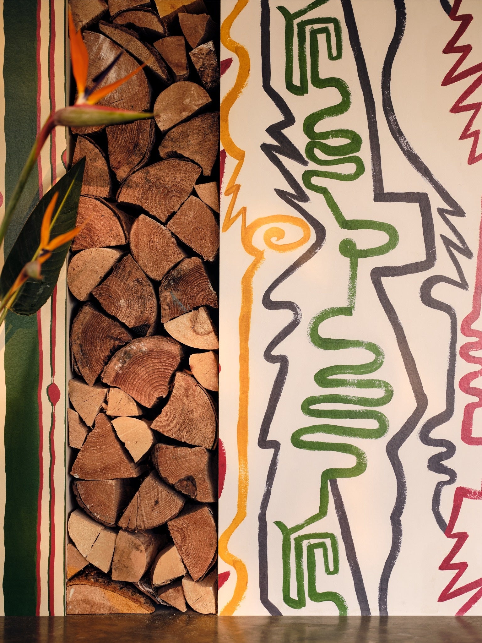 Close-up of stacked firewood next to bold abstract line art wallpaper in green, yellow, and red with playful, wavy shapes and outlines.