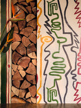 Close-up of stacked firewood next to bold abstract line art wallpaper in green, yellow, and red with playful, wavy shapes and outlines.
