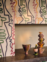 Colourful abstract line art wallpaper with red, green, yellow, and black face outlines framing a fireplace, with handmade ceramic vases on the mantle.