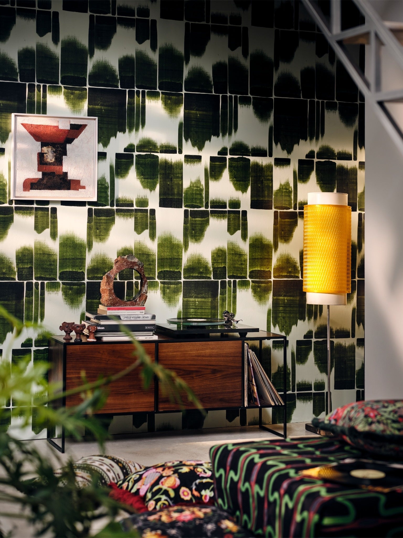Green graphic wallpaper with black pattern behind a mid-century sideboard, art print, yellow wall lamp, and vinyl records in a cozy music room.