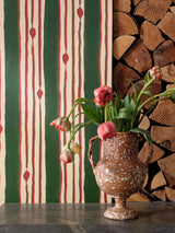 Tulip-filled rustic vase in front of green and red vertical striped wallpaper and stacked firewood, creating a warm, earthy atmosphere.
