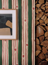 Framed abstract art hanging over green and red vertical striped wallpaper beside a stack of firewood in a modern eclectic interior.