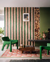 Green and red vertical striped wallpaper behind stacked firewood, green chairs, a low wood table, and a geometric rug in a modern lounge.