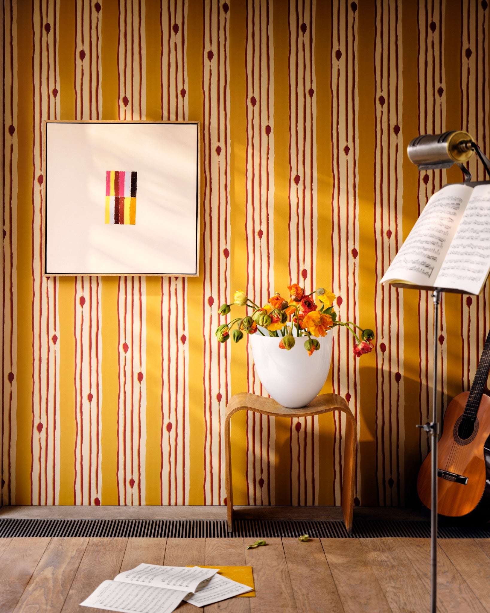 Yellow striped wallpaper with red dots behind a wooden chair, white vase of poppies, framed abstract art, guitar, and scattered sheet music.