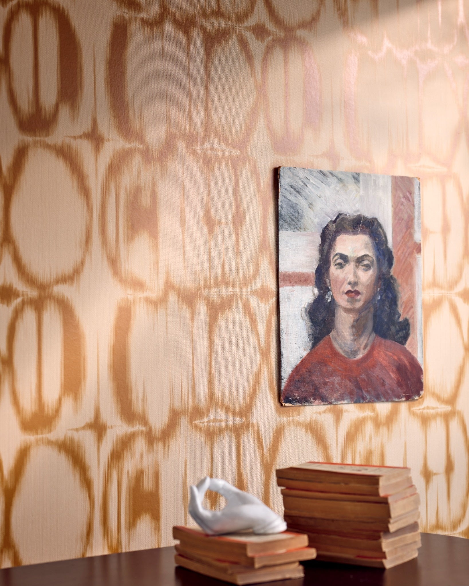 Beige and tan ikat-style patterned wallpaper behind a small portrait painting of a woman, with a stack of vintage books and a white sculpture.