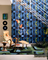 Blue, black, and green geometric patterned wallpaper behind a wood lounge chair, orange floor lamp, and a bold abstract patterned rug.