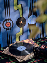 Close-up of blue wallpaper with abstract black, red, and green shapes, showing a vinyl record and vintage album covers resting on a patterned bench.