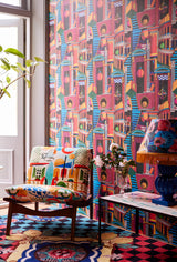 Vibrant room with bold geometric PIAZZA DEGLI ARLECCHINI wallpaper, colourful upholstered chair, patterned rug, floral lamp, and a modern glass table.