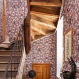 A rustic wooden staircase with floral wallpaper in deep red and brown hues. The space features vintage decor, framed artwork, and warm wooden architectural details