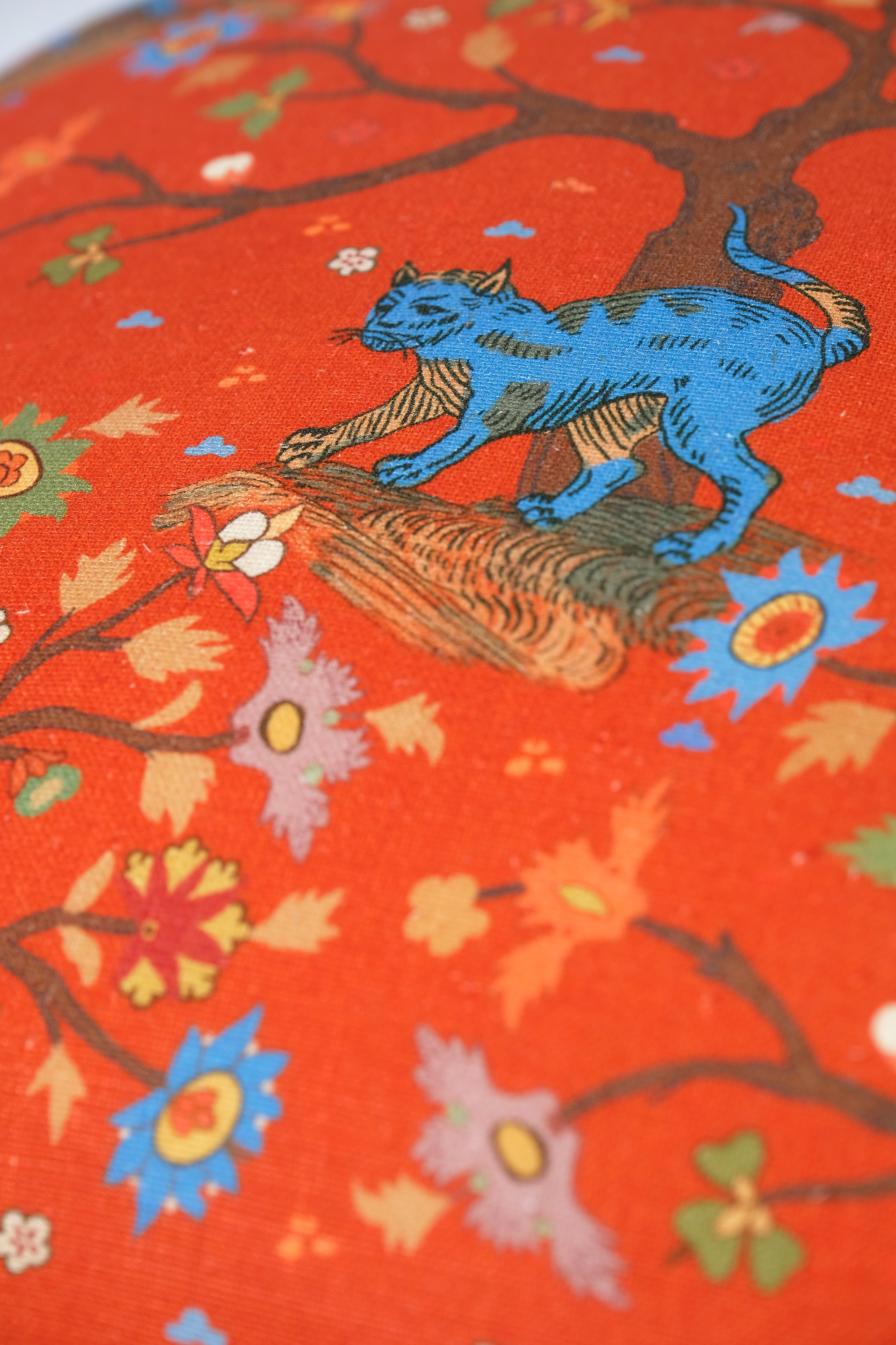 A close-up of a vibrant orange textile featuring a whimsical illustration of a blue cat walking on a tree branch with stylized flowers and leaves.