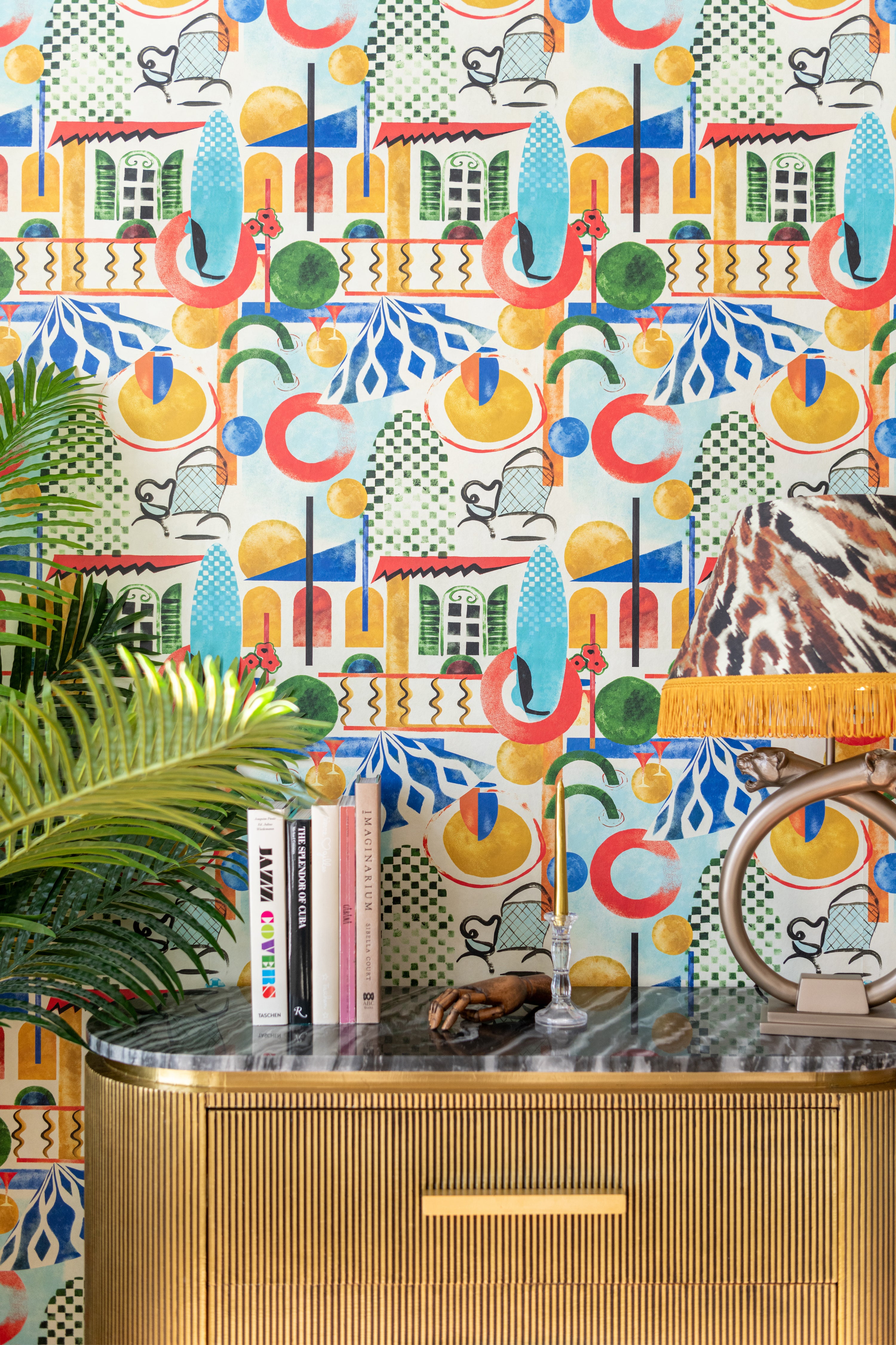 A vibrant space with VERANDA wallpaper, a gold-ribbed sideboard, books, a palm plant, and a fringed leopard-print lampshade.