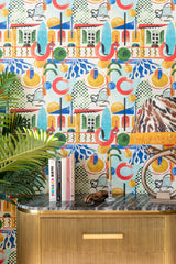 A vibrant space with VERANDA wallpaper, a gold-ribbed sideboard, books, a palm plant, and a fringed leopard-print lampshade.