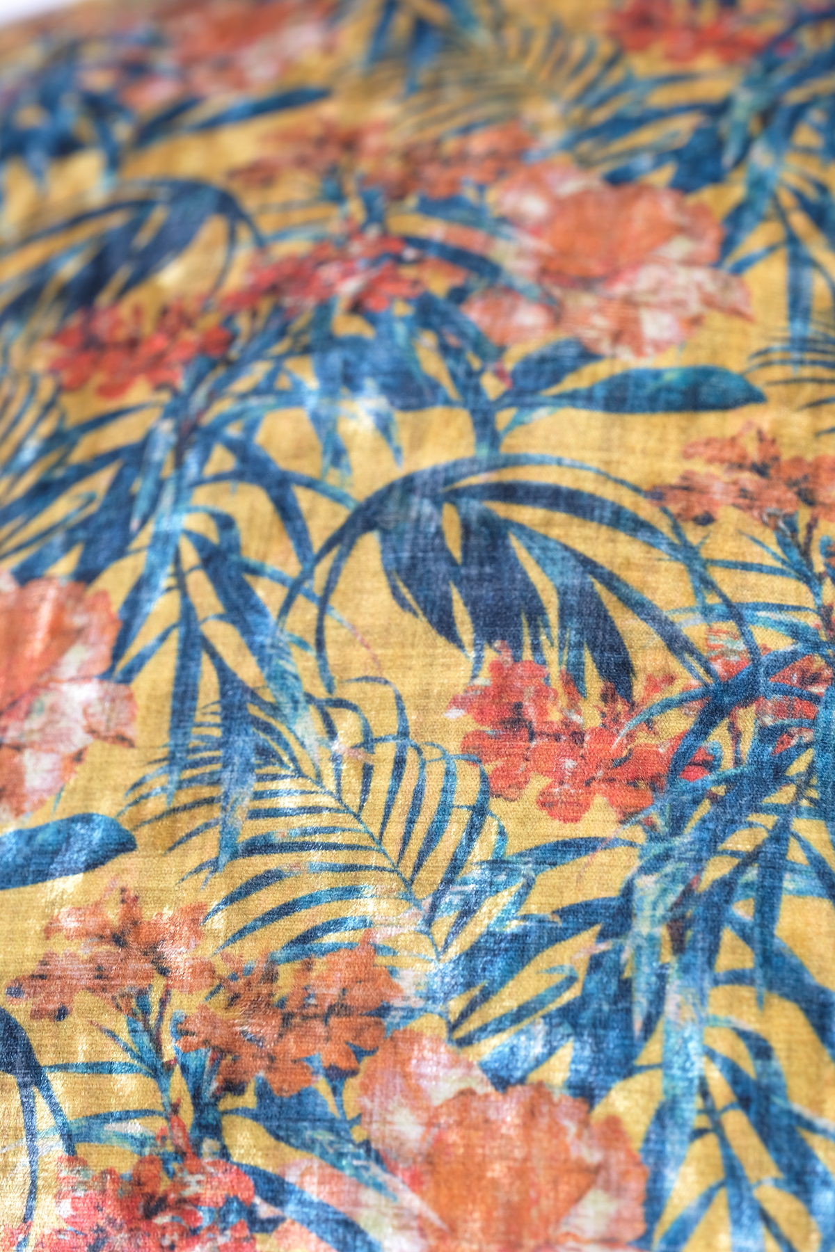 A close-up of a tropical fabric print featuring blue palm leaves and coral-orange flowers on a mustard yellow background.