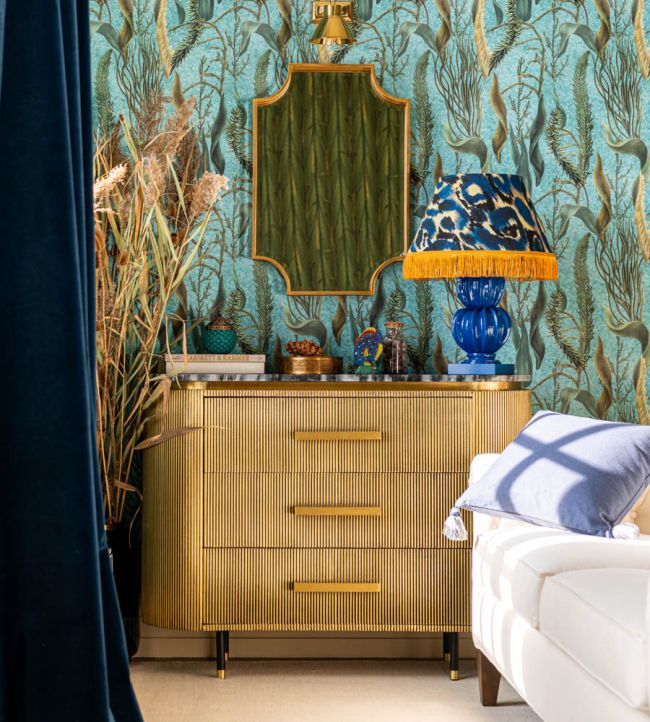 A luxurious bedroom with aquatic-inspired wallpaper, a gold dresser, and bold blue and gold decor. A plush white couch and natural light add elegance.