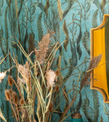 A close-up of an aquatic-themed wallpaper in deep blue-green hues, featuring underwater plants. Dried grasses and a gold mirror add warmth and texture.