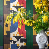 Colourful geometric wallpaper with red, green, blue, yellow, and black shapes, behind a white vase filled with yellow flowers