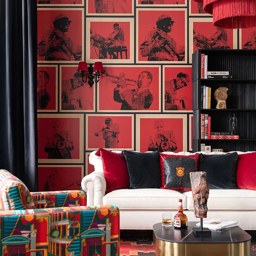 A living room with a red jazz musician wallpaper, white sofa with red and black cushions, black bookcase, and vibrant patterned armchair.