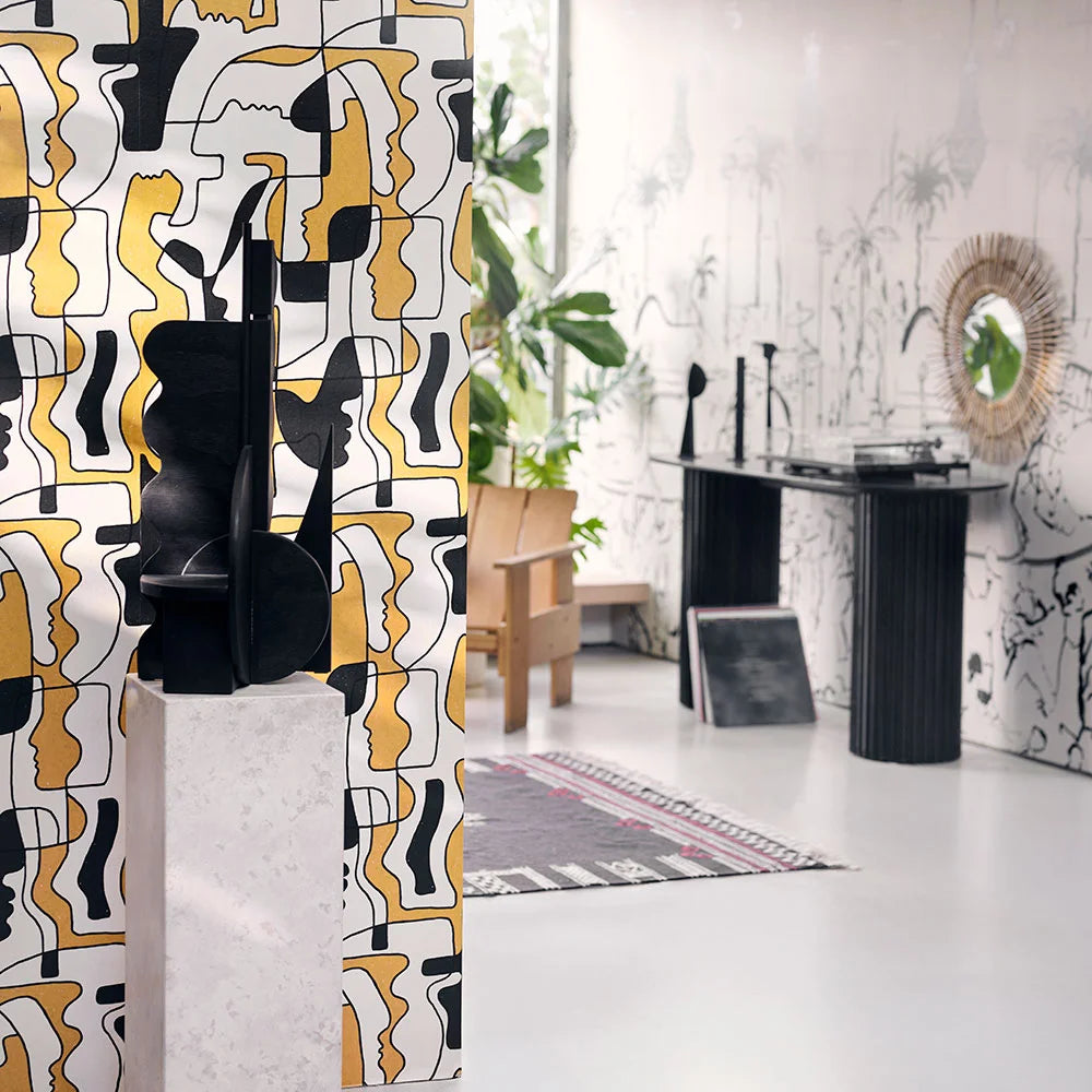 A modern interior featuring gold and black abstract face wallpaper on a partition, with sculptural decor and a sleek black console table in the background.